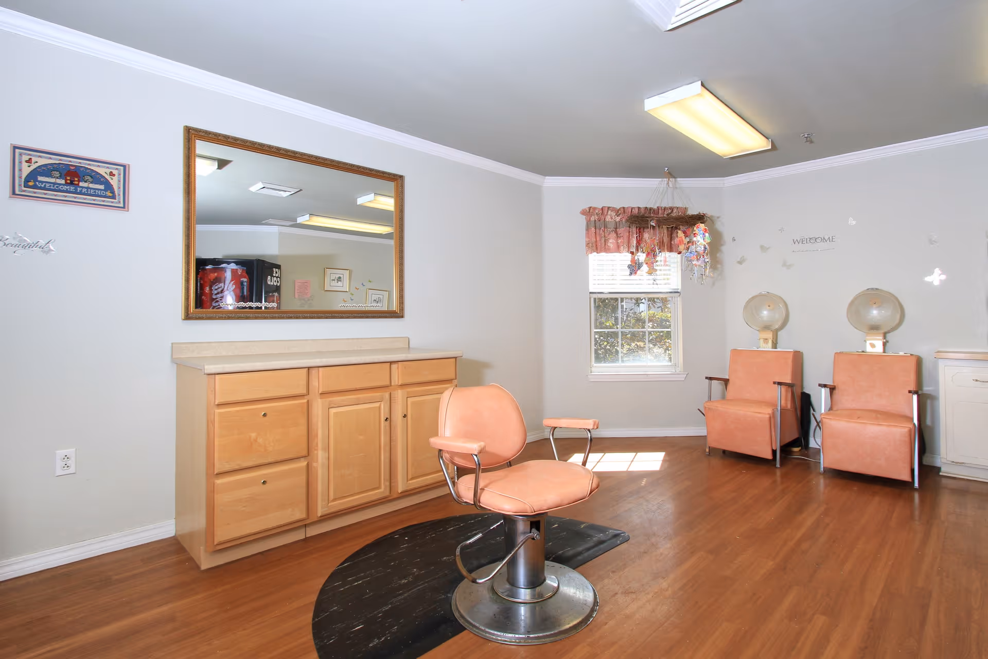 Interior view of a hair salon area in an assisted living facility with a salon chair in the center on a black mat, a wooden cabinet with drawers and a large mirror above it on the left wall, two peach-colored salon chairs with hair dryers attached on the right side near a window with floral curtains, and light wooden flooring.