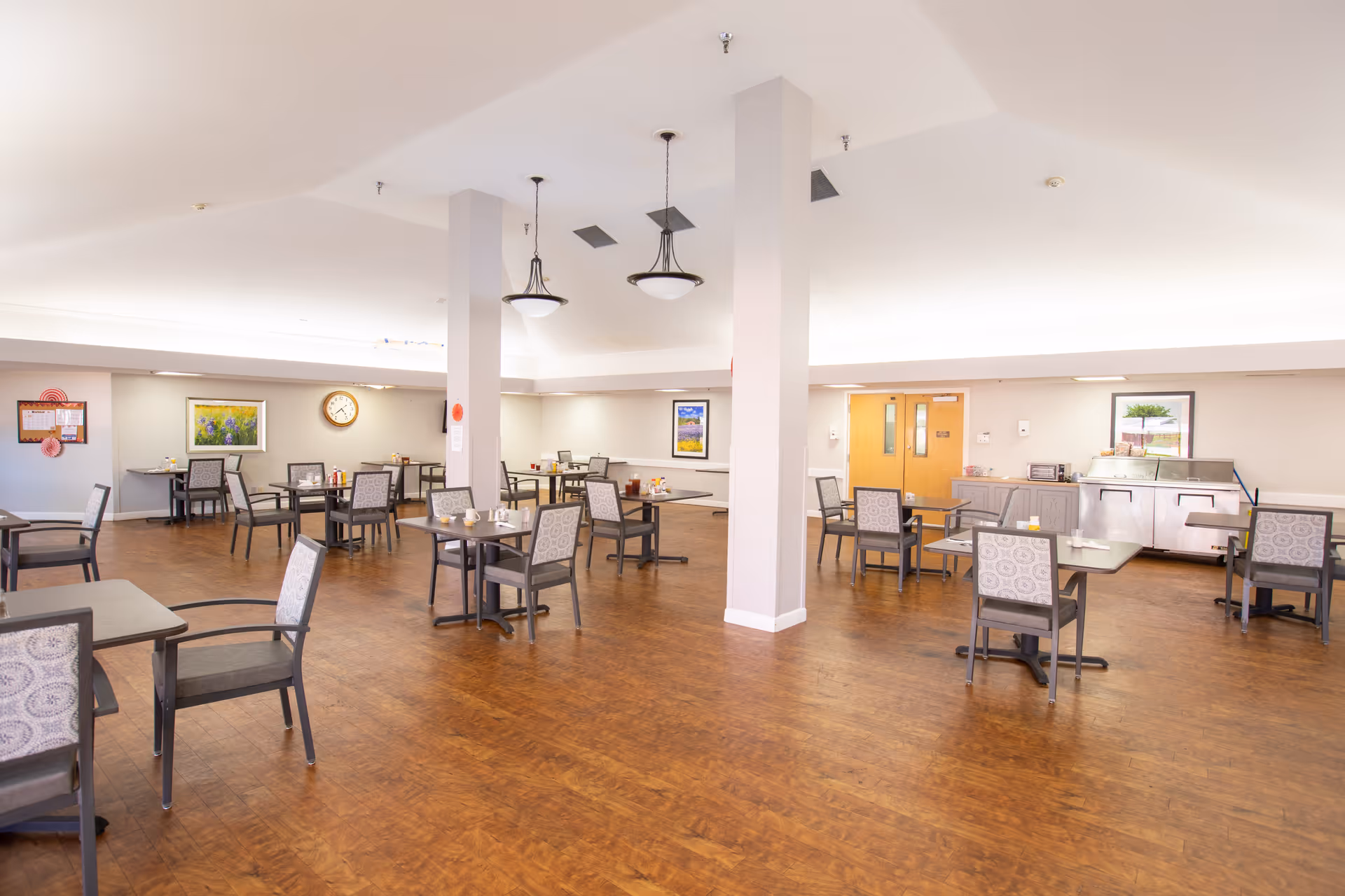 A spacious dining room with multiple tables and chairs arranged neatly on a wooden floor. The room is well-lit with ceiling lights and has light-colored walls adorned with framed artwork and a clock. There is a serving area with a counter and some appliances near the back wall.