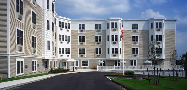 Exterior view of a multi-story senior living facility building with beige and white siding, multiple windows, a paved driveway, green lawn areas, and an American flag on a flagpole in front.