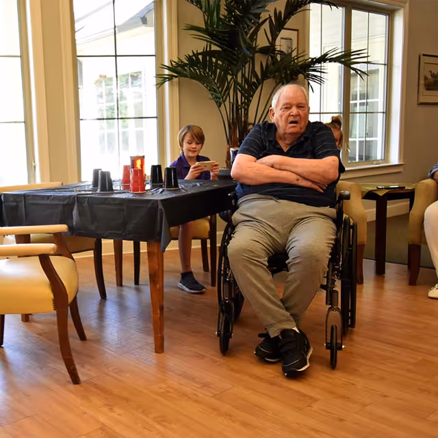 An elderly man sitting in a wheelchair with arms crossed in a room with wooden flooring and large windows. Behind him, a young boy is seated at a table covered with a black tablecloth, playing on a handheld device. The room has beige chairs and a large potted plant near the windows.