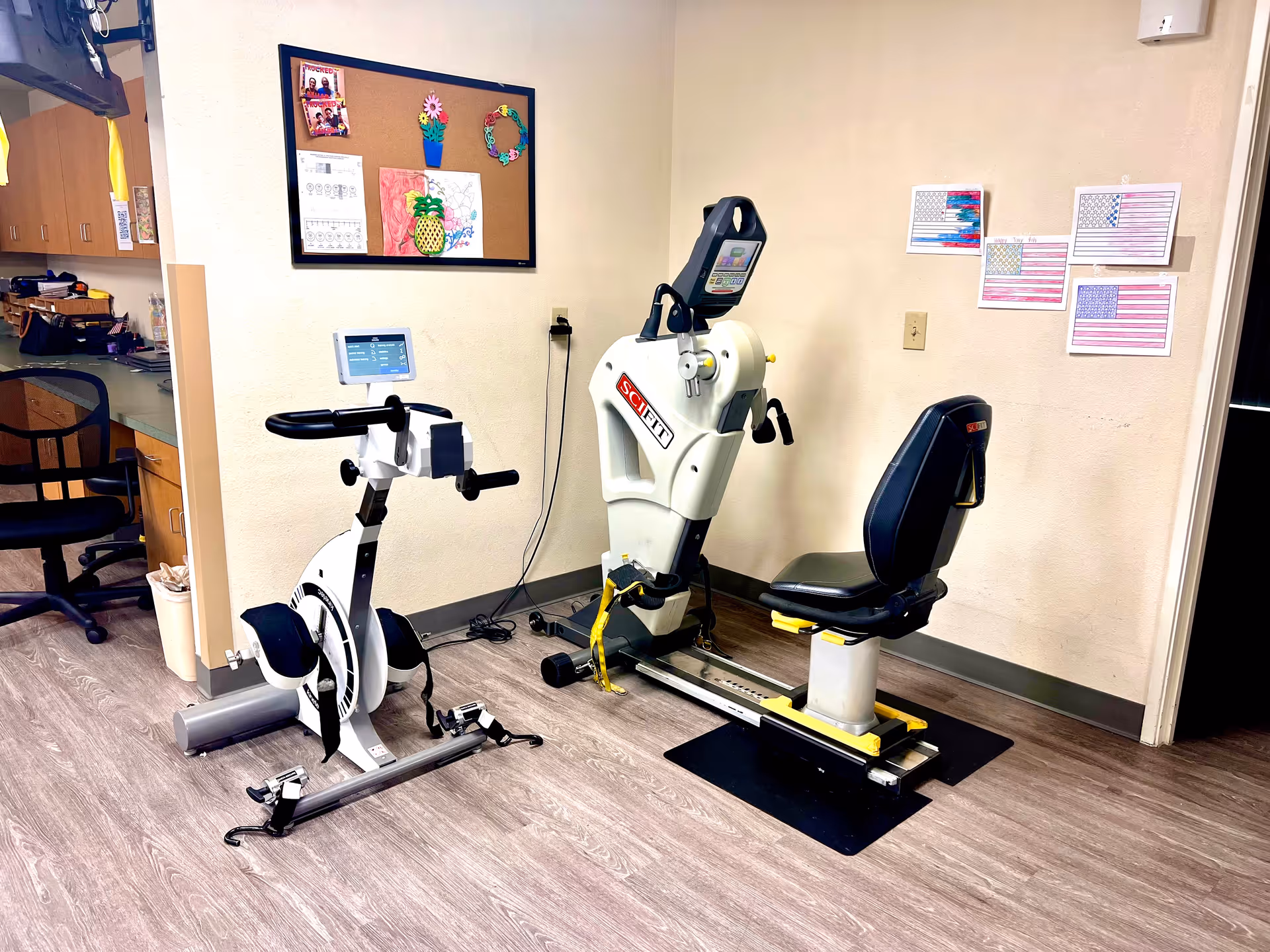 A small exercise room with two pieces of fitness equipment, including a stationary bike and a seated exercise machine. The room has light-colored walls with children's artwork and American flag drawings pinned on the wall. There is a bulletin board with colorful decorations and a desk area with a chair and cabinets on the left side.