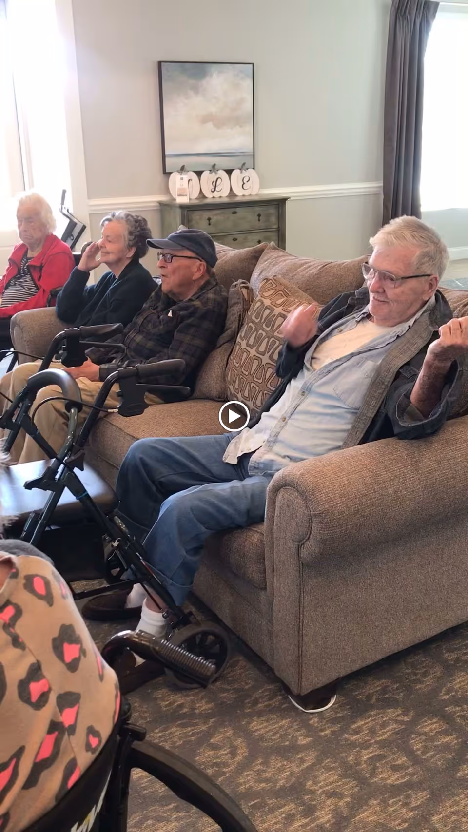 A group of elderly people sitting on a couch and chairs in a living room area, some using walkers. The room has neutral-colored walls, a painting, and decorative items on a small dresser behind the couch.