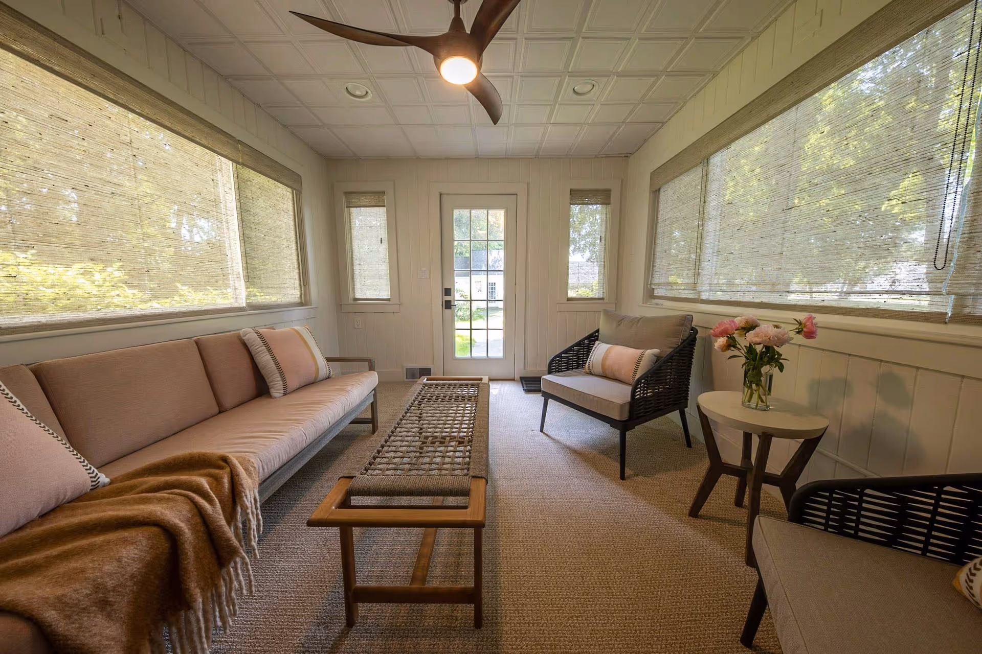 Bright sunroom-style living area with a long sofa, wicker chairs, a woven bench, side table with flowers, large windows, and a door to the outside.