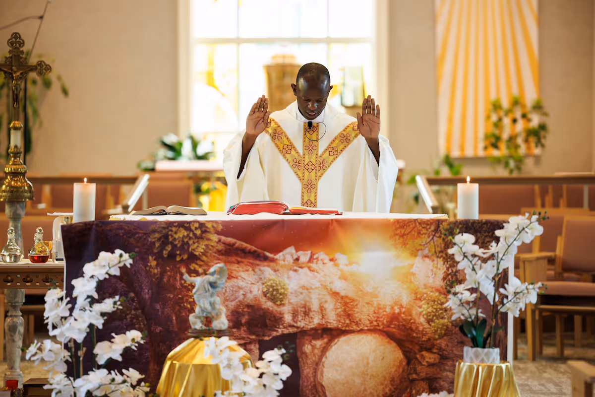 A priest in white and gold vestments stands with raised hands behind an altar decorated with flowers, candles, and a religious cloth in a chapel.