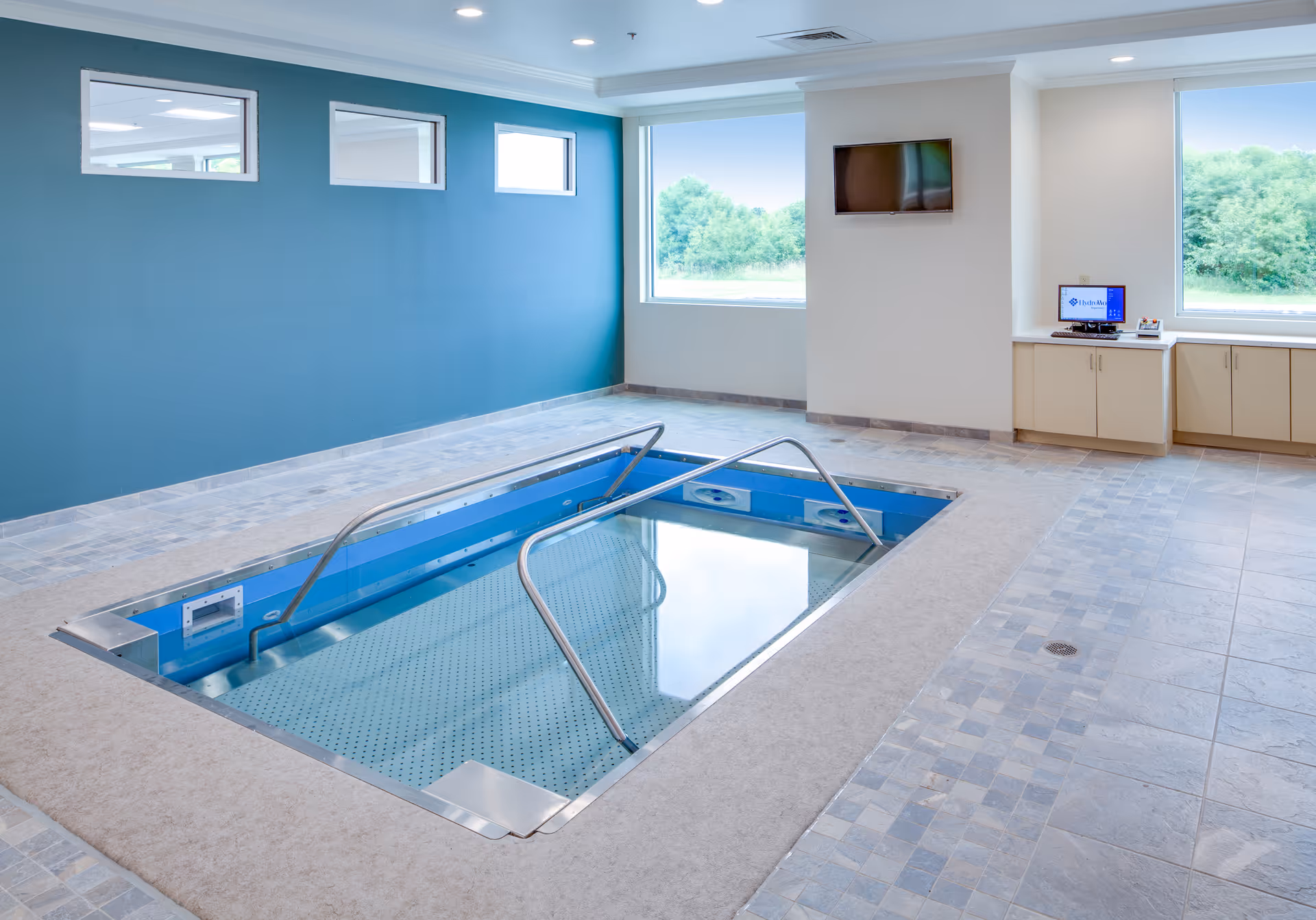 Indoor therapy pool with stainless steel handrails in a bright room featuring blue and white walls, tiled floor, two large windows showing greenery outside, a wall-mounted TV, and a computer workstation on a cabinet.