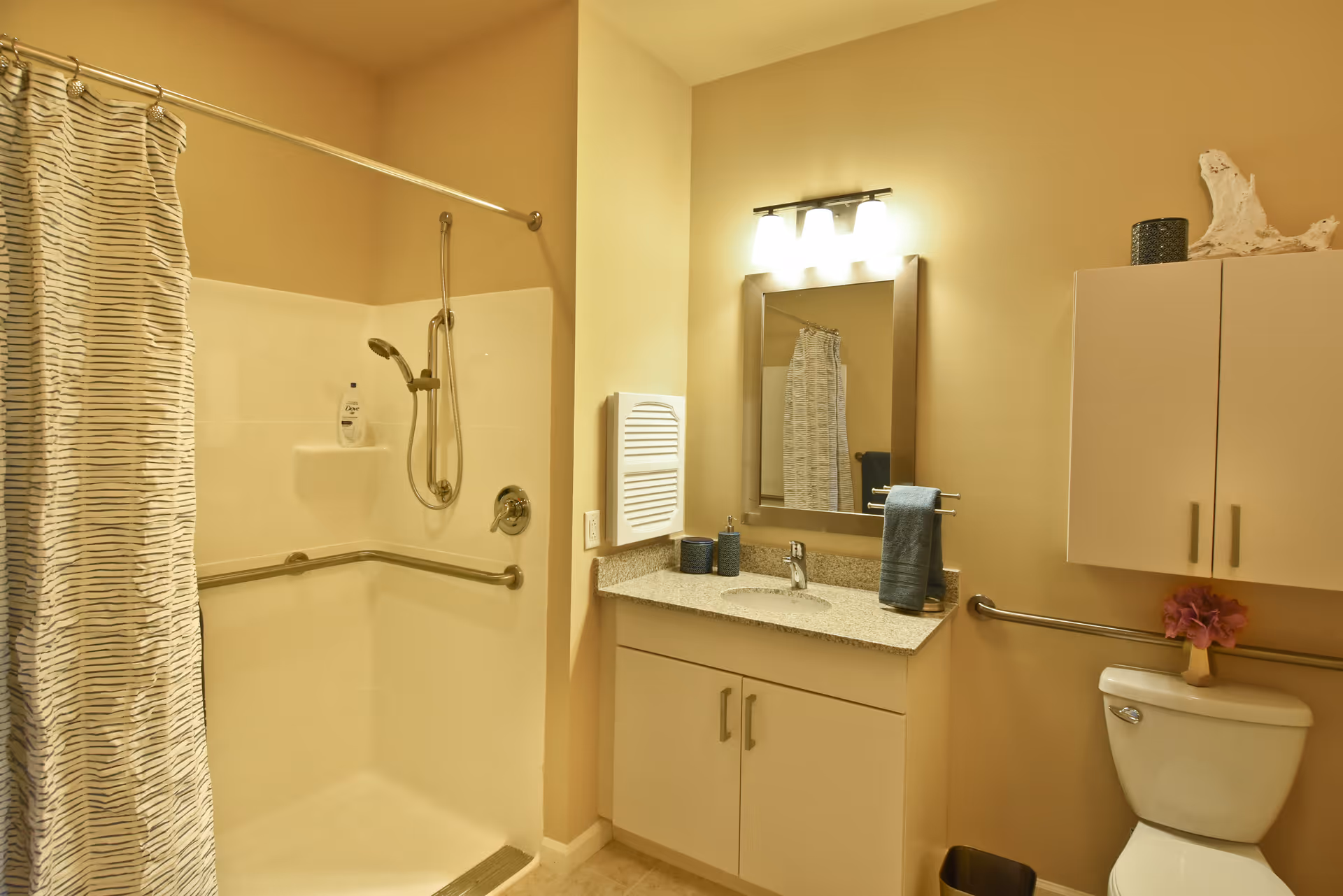 A bathroom with a walk-in shower featuring a handheld showerhead and a striped shower curtain. There is a vanity with a granite countertop, a sink, and a mirror with three lights above it. A cabinet is mounted on the wall above the toilet, which has a small vase with pink flowers on top. The bathroom has beige walls and safety grab bars near the shower and toilet.