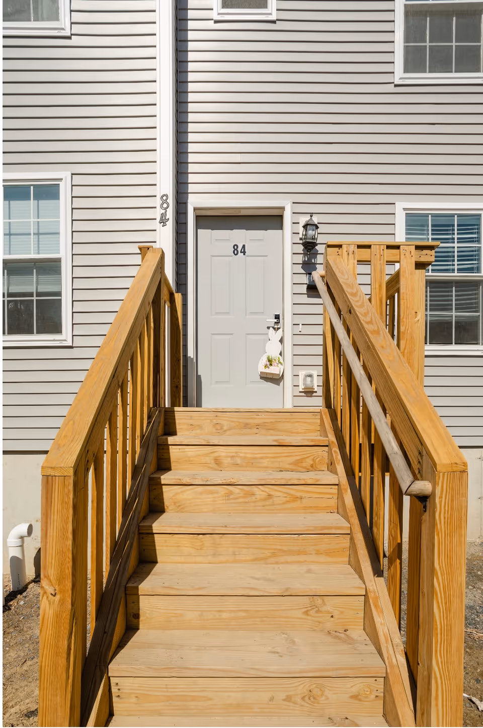 Wooden stairs with railings leading up to a white door numbered 84 on the exterior of a beige siding building with windows on either side.