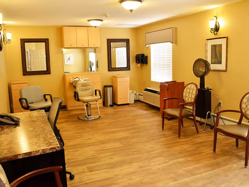 Interior room with wooden floor and yellow walls featuring salon chairs, mirrors, a hair dryer, and several armchairs arranged around the room, suggesting a hair salon or beauty care area.