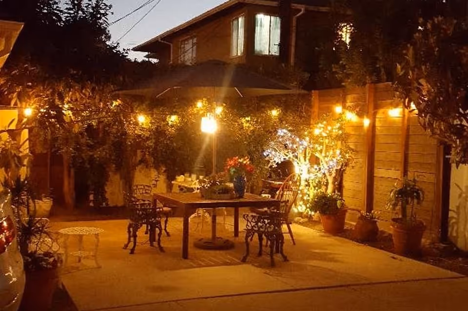 Outdoor patio area at night with string lights hanging above a wooden table surrounded by chairs. Potted plants and small trees decorated with lights are placed around the patio, creating a warm and inviting atmosphere. A house with lit windows is visible in the background.