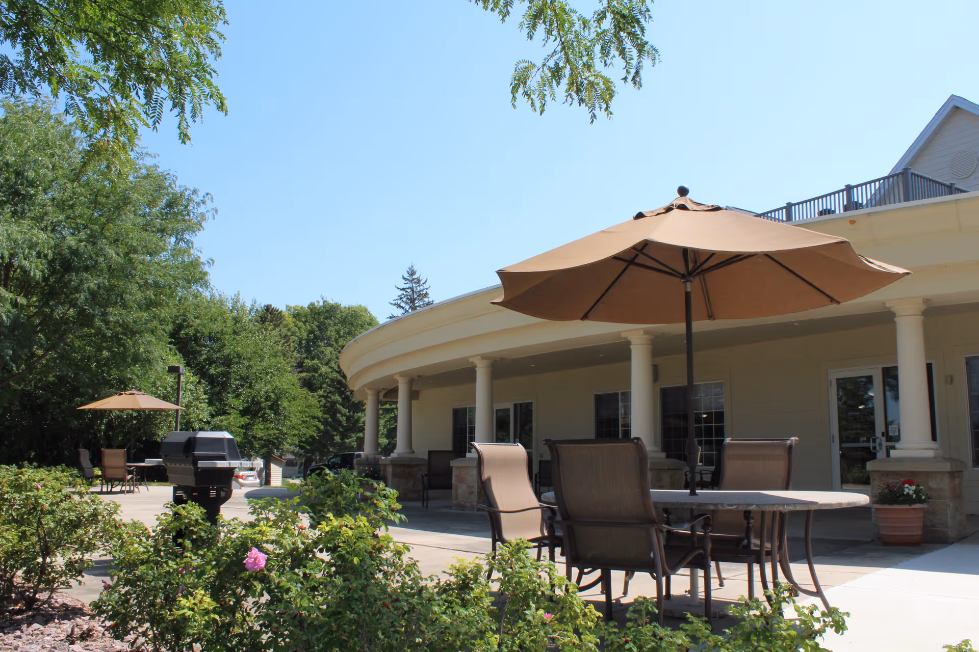 Outdoor patio area at Vista Prairie at Ridgeway on 23rd with tables and chairs under large brown umbrellas, surrounded by greenery and flowers, adjacent to a building with columns and windows.