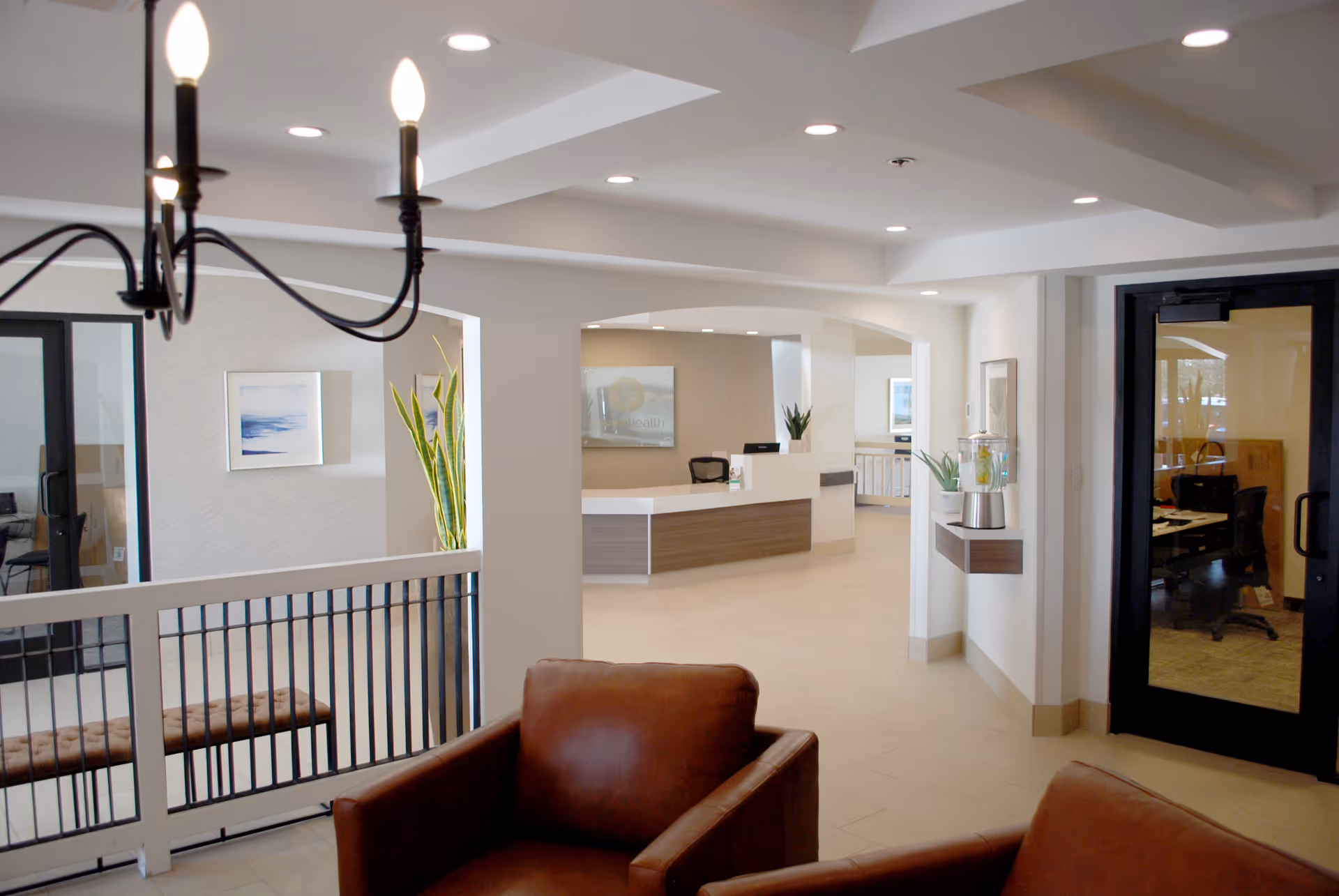 Interior view of a senior living facility reception area with a modern front desk, leather armchairs in the foreground, a chandelier hanging from the ceiling, and glass doors leading to office spaces. The space is well-lit with recessed lighting and decorated with plants and framed artwork.