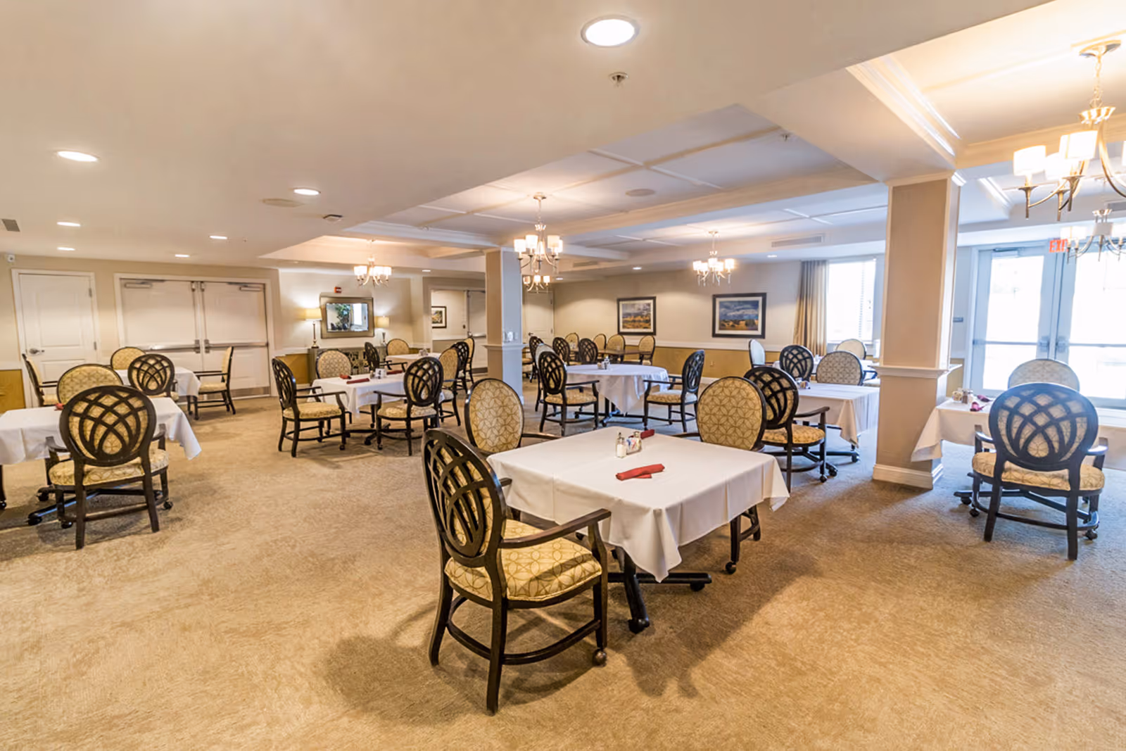 A spacious dining room with multiple tables covered in white tablecloths, each surrounded by cushioned chairs with patterned upholstery. The room is well-lit with ceiling lights and chandeliers, and there are framed pictures on the walls. Large windows and glass doors allow natural light to enter the room.