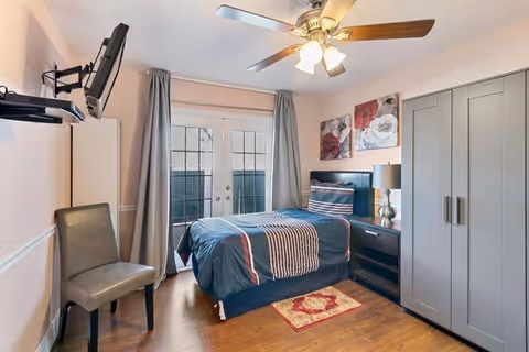 A neatly arranged bedroom with a single bed covered in a blue and striped comforter, a bedside table with a lamp, a gray wardrobe, a wall-mounted TV, a gray chair, and French doors with curtains leading outside. The room has wooden flooring, a ceiling fan with lights, and two floral paintings on the wall above the bed.