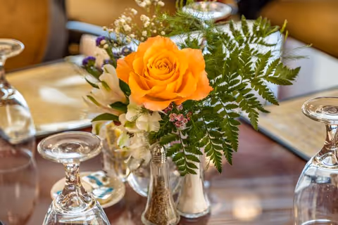 Close-up of a dining table centerpiece featuring an orange rose surrounded by greenery and small white flowers, with upside-down wine glasses and salt and pepper shakers on a polished wooden table.
