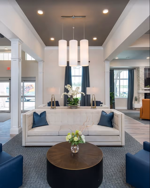 A modern living room area in a senior living facility featuring a beige tufted sofa with two dark blue pillows, a round wooden coffee table with a small floral arrangement, two table lamps with white shades behind the sofa, and three cylindrical pendant lights hanging from the ceiling. Large windows with dark curtains and additional seating are visible in the background.