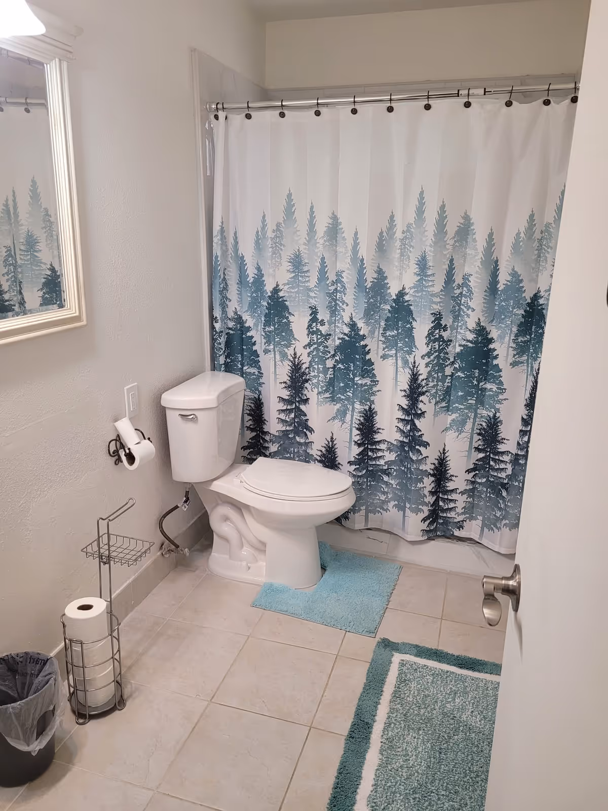 A clean bathroom with a white toilet, a shower curtain featuring a forest tree design in shades of blue and green, a small blue bath mat in front of the toilet, a larger blue and white rug on the tiled floor, a wall-mounted mirror, a toilet paper holder with a roll, a metal stand holding extra toilet paper rolls, and a small trash can.