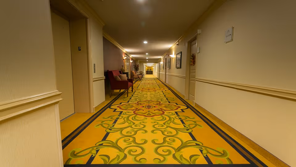 A long, well-lit hallway in a senior living facility with patterned yellow and green carpet, beige walls with chair rails, several doors on each side, and a few chairs and framed pictures along the walls.