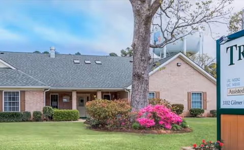 Exterior view of Trinity Timbers Assisted Living building with lawn, trees, and flowering bushes.