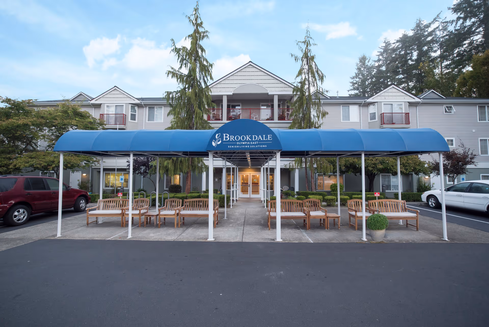 Front exterior view of Brookdale Olympia East senior living facility with a blue canopy entrance, wooden benches, and parked cars on either side.