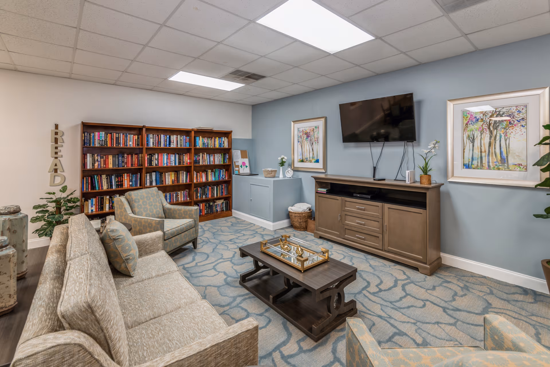 A cozy living room area with a beige sofa and armchair, a wooden coffee table with decorative items, a wooden bookshelf filled with books, a TV mounted on a light blue wall, and two framed colorful paintings. The room has a patterned carpet and a ceiling with fluorescent lights.