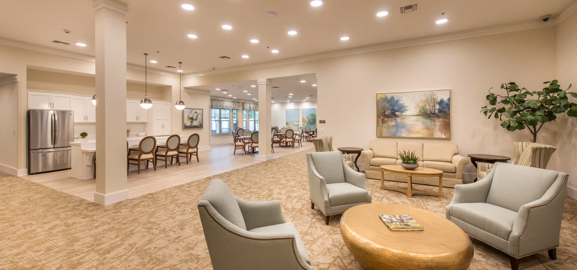 A spacious and well-lit common area in a senior living facility featuring a seating area with three armchairs and a sofa around a round coffee table. The room has beige walls, carpeted flooring, and decorative paintings on the walls. In the background, there is a dining area with tables and chairs, and a kitchen area with a stainless steel refrigerator and a counter with bar stools.