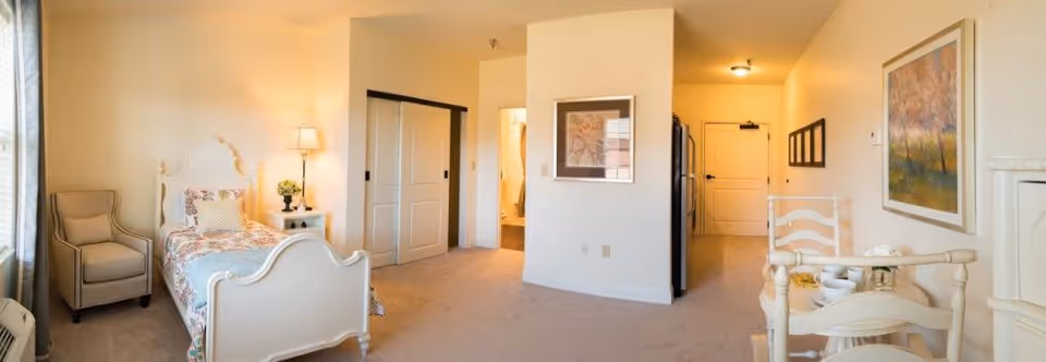 A bright and cozy senior living apartment featuring a single bed with a floral bedspread, a beige armchair, a nightstand with a lamp and flowers, a small dining table with two chairs, and framed artwork on the walls. The room has beige carpeting and light-colored walls, with a doorway leading to a bathroom and a kitchen area with a refrigerator.