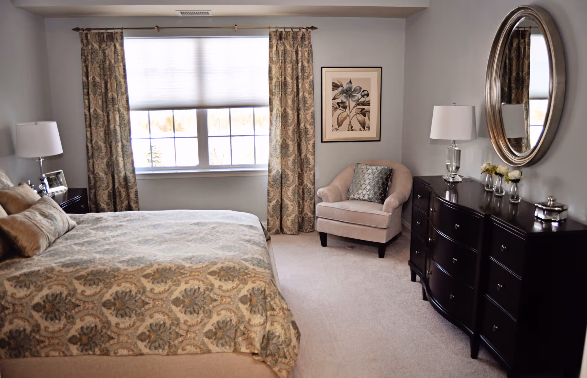 A cozy bedroom with a large bed covered in a patterned bedspread, a beige armchair with a decorative pillow, a dark wooden dresser with a round mirror above it, two table lamps, and a window with patterned curtains.