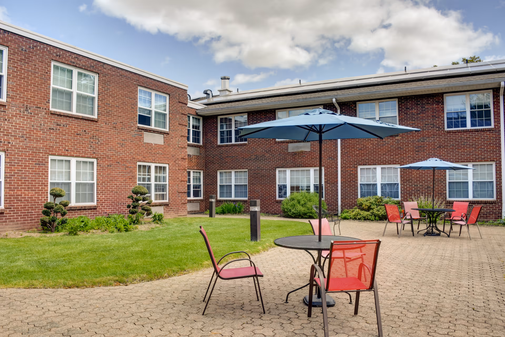 Outdoor patio area at The Court of Saint James featuring round tables with blue umbrellas and red chairs on a paved surface, surrounded by a well-maintained lawn and a two-story brick building with multiple windows under a partly cloudy sky.