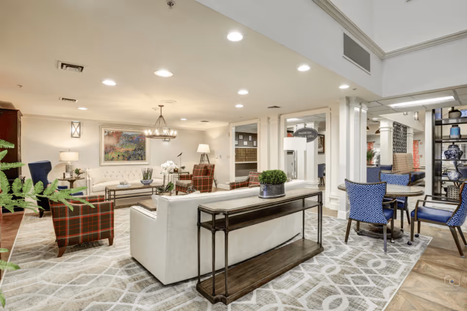 A spacious and well-lit living room area in a senior living facility featuring a white sofa, plaid armchairs, a coffee table, and a side table with a lamp. The room has a patterned carpet, recessed ceiling lights, and decorative plants. In the background, there is a dining area with blue cushioned chairs and round tables, separated by white columns and a shelving unit with decorative items.