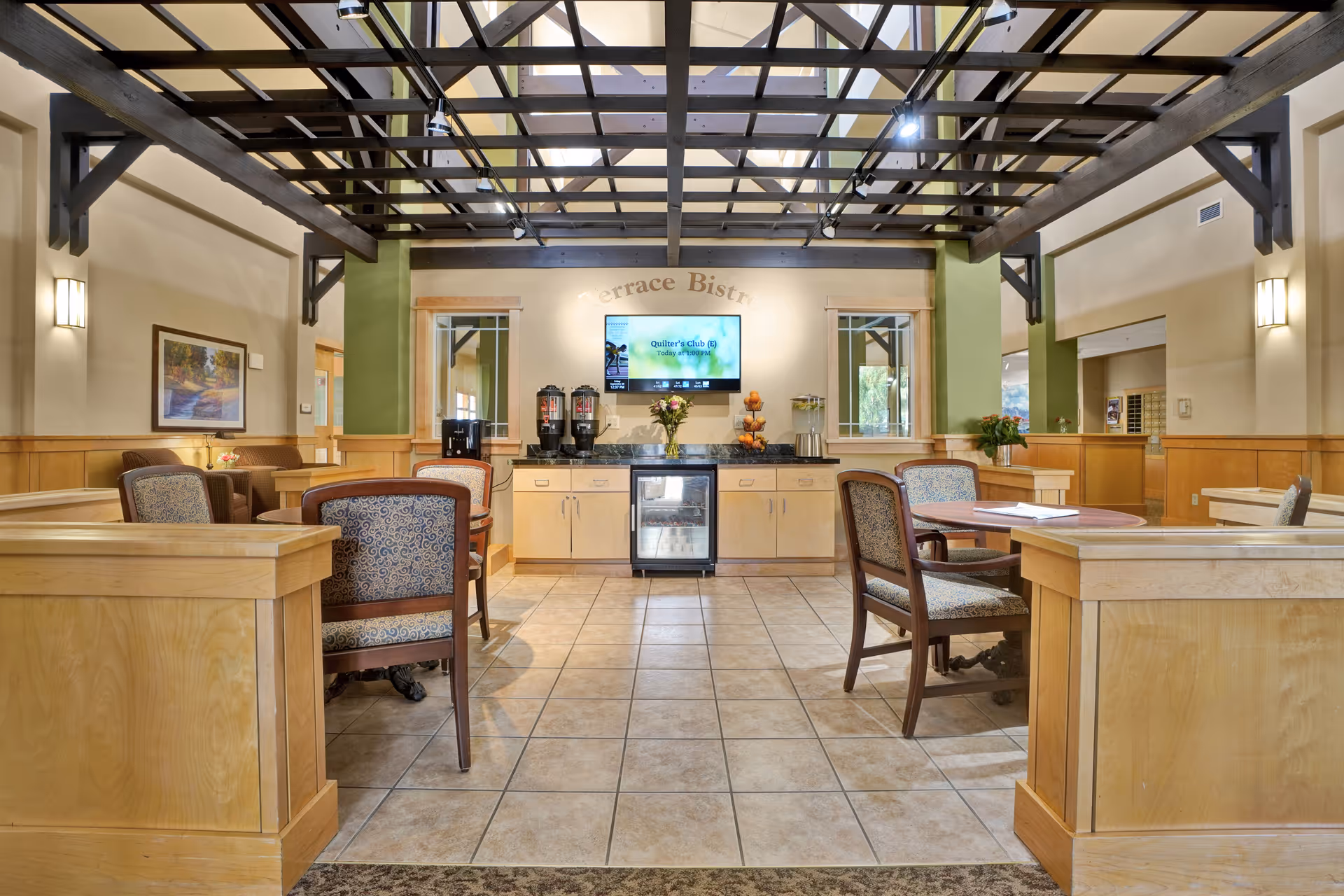 Interior view of a senior living facility's dining or common area with wooden tables and chairs, a tiled floor, and a beverage station against the far wall under a ceiling with exposed beams and skylights. The walls are painted in neutral tones with green accents, and there are framed pictures and wall sconces providing lighting.