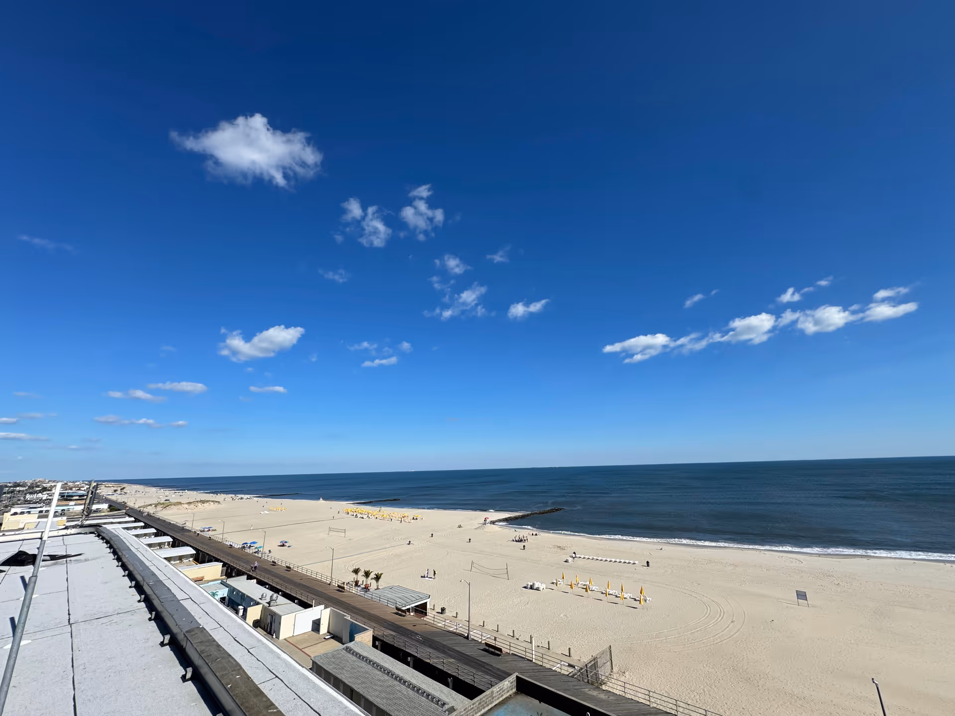View of a sandy beach with a few people scattered along the shore, a calm ocean under a blue sky with scattered clouds, and a boardwalk running parallel to the beach with buildings and beach facilities visible.