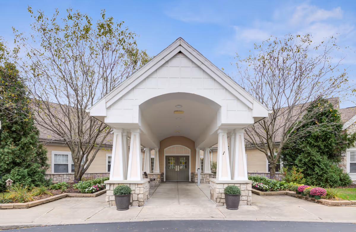 Entrance to a senior living community building with a covered driveway supported by white pillars. The building has beige siding and stone accents, with landscaped flower beds and trees on either side of the entrance.