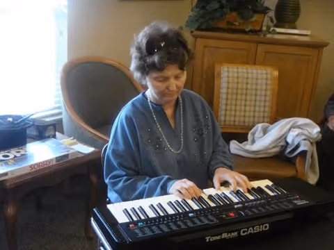 An elderly woman wearing a blue sweater is playing a Casio Tone Bank keyboard in a cozy room with a chair, a wooden cabinet, and a table with board games on it.