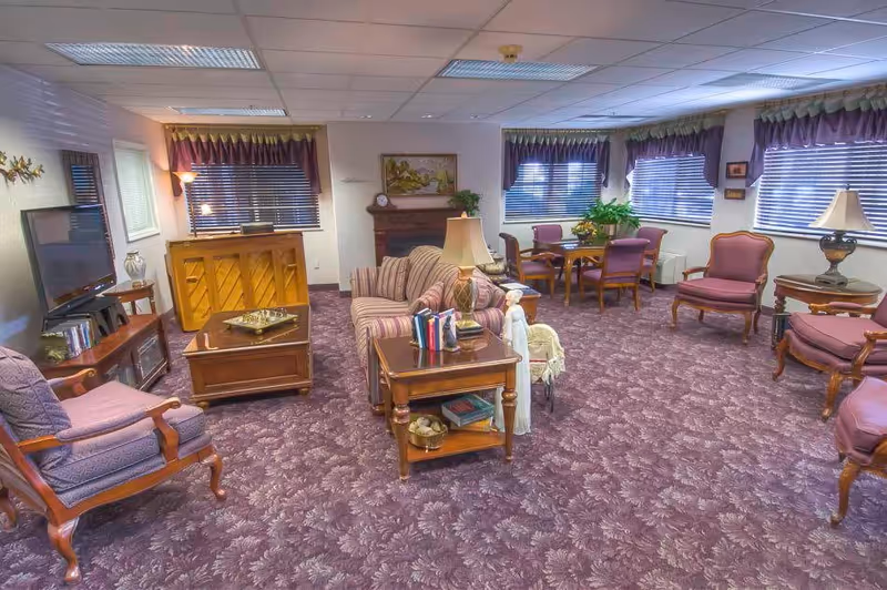 A spacious living room with purple patterned carpet, multiple upholstered chairs and sofas, wooden tables with lamps and books, a piano, a flat-screen TV on a wooden stand, and several windows with blinds and valances.