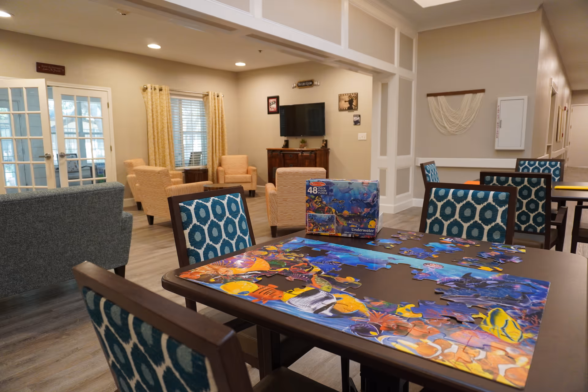 A common area in a senior living facility featuring a table with a partially completed colorful underwater-themed puzzle and its box. The room has patterned chairs around the table, beige armchairs, a TV mounted on the wall, and large windows with curtains letting in natural light.