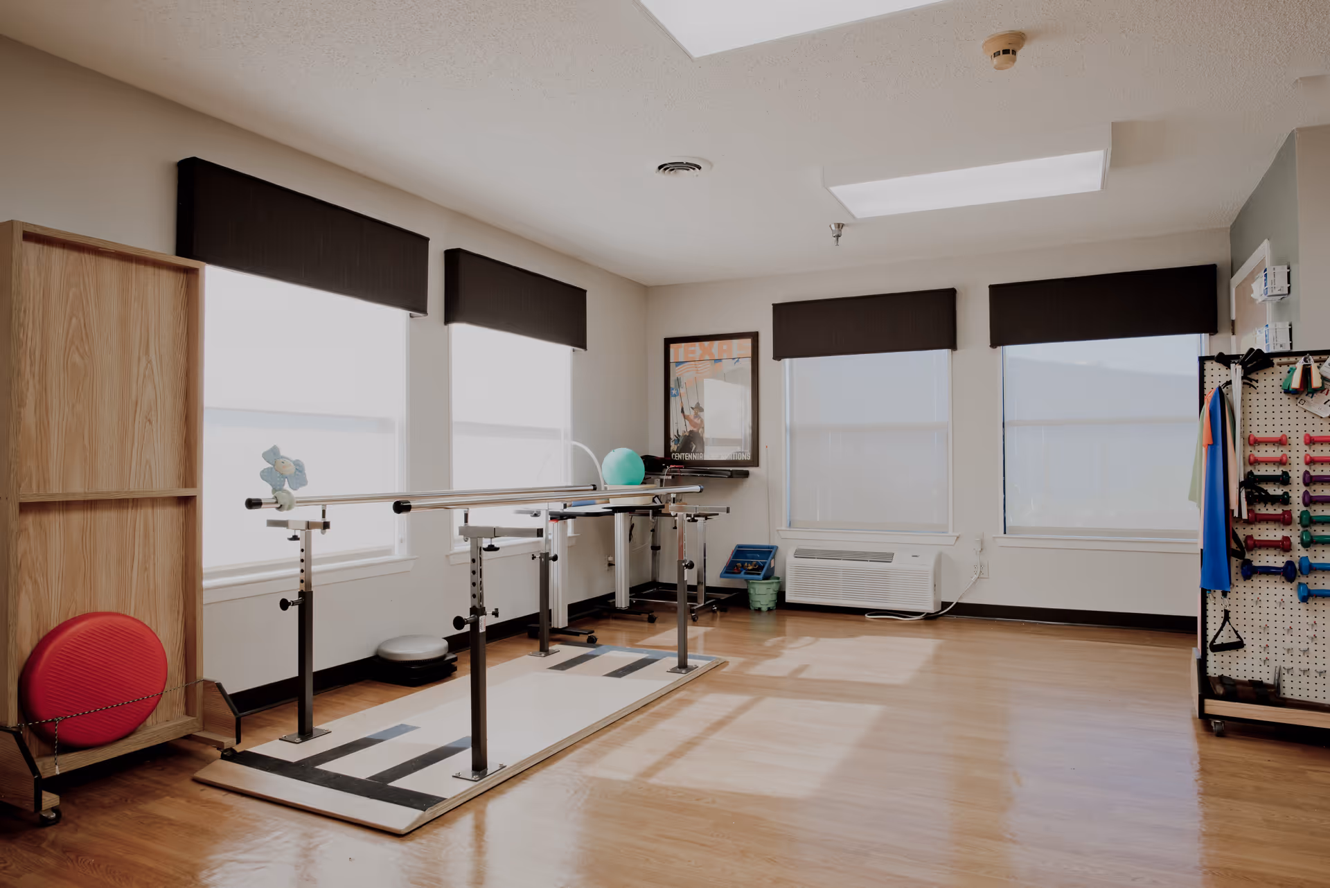 A bright rehabilitation room with wooden floors and large windows covered with dark valances. The room contains parallel bars for physical therapy, a red exercise ball on a wooden stand, a green exercise ball on a table, and a pegboard with various colorful exercise equipment such as weights and resistance bands. There is also an air conditioning unit below the windows and a framed poster on the wall.