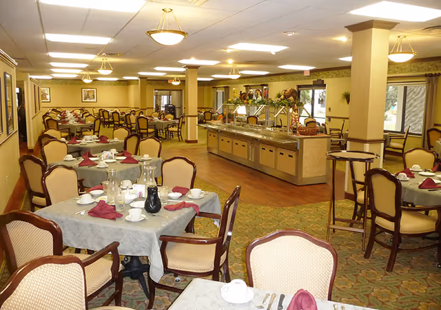 A spacious dining room in a retirement center with multiple tables covered in gray tablecloths, each set with white cups, saucers, silverware, and burgundy napkins. There is a buffet area with food warmers and decorative plants in the background. The room has beige walls, carpeted and wooden flooring, and several ceiling lights.