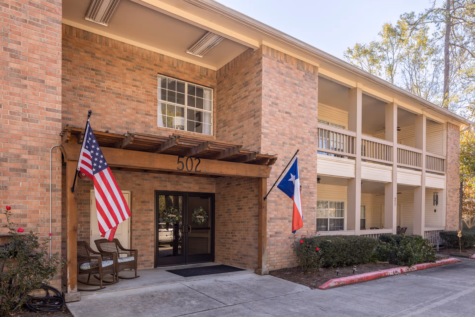 Exterior view of a brick building with the number 502 above the entrance. Two flags, an American flag and a Texas state flag, are mounted on either side of the entrance. There are two wicker chairs with cushions on the porch area. The building has two stories with balconies and some landscaping with bushes and flowers near the entrance.