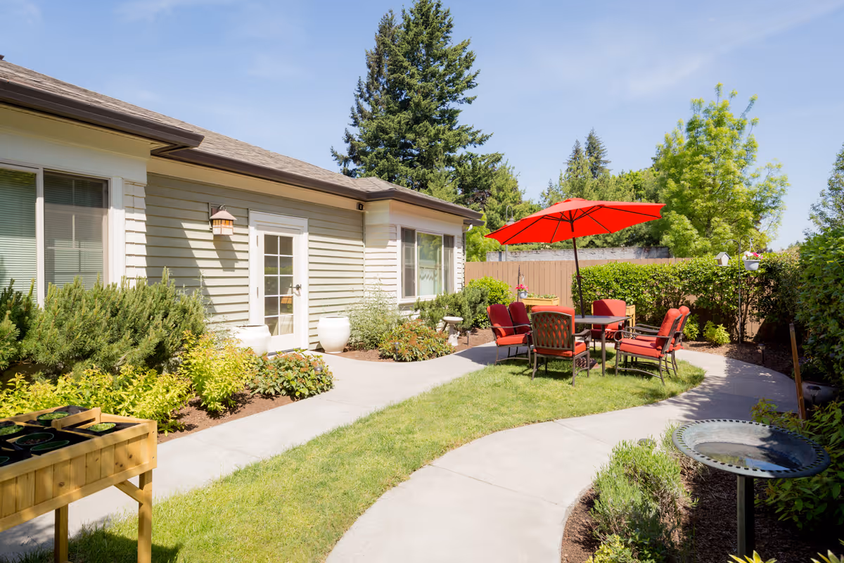 Outdoor patio area at Brookdale Vancouver Orchards with a curved concrete pathway, green grass, and a seating area featuring a table with a red umbrella and red cushioned chairs. The patio is surrounded by bushes, plants, and trees under a clear blue sky.