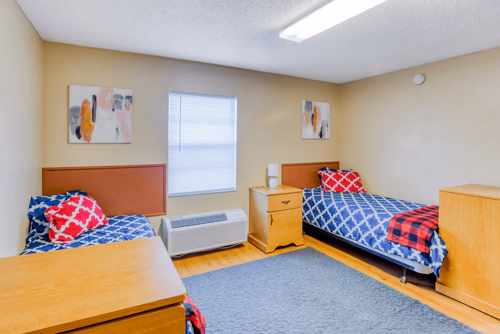 A simple bedroom with two single beds, each with blue and white patterned bedding and a red pillow. There is a wooden nightstand with a small lamp between the beds, a window with blinds, two abstract paintings on the walls, and a blue rug on the wooden floor.