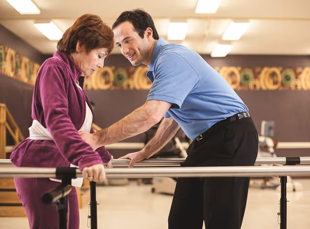 An elderly woman in a purple tracksuit is using parallel bars for physical therapy while a male therapist in a blue shirt assists and supports her in a rehabilitation room.