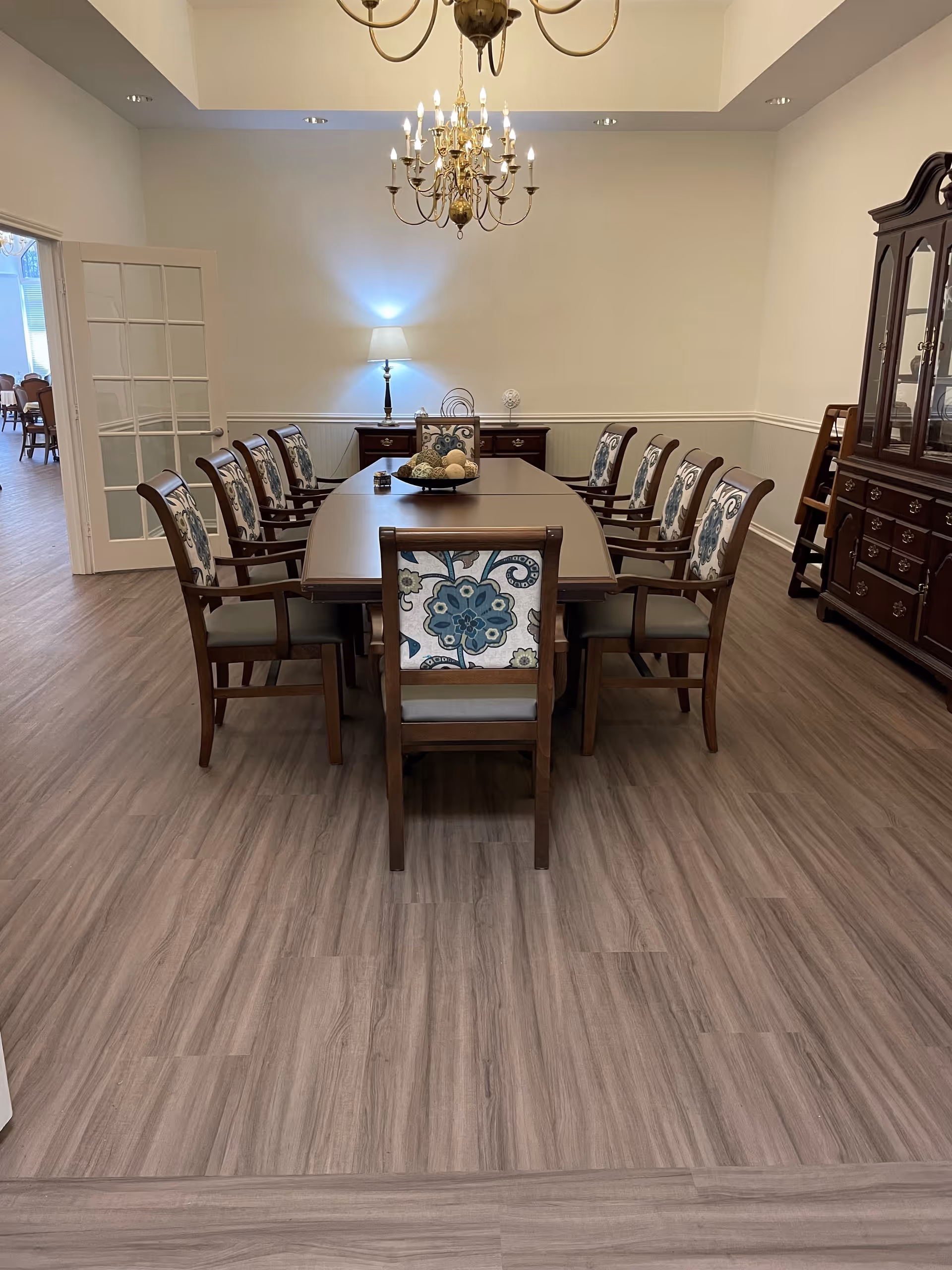 A formal dining room with a long rectangular wooden table surrounded by ten wooden chairs with floral patterned upholstery. A chandelier hangs above the table, and a sideboard with a lamp and decorative items is against the far wall. The floor is wood laminate, and a china cabinet is visible on the right side. French doors lead to another room with additional seating.