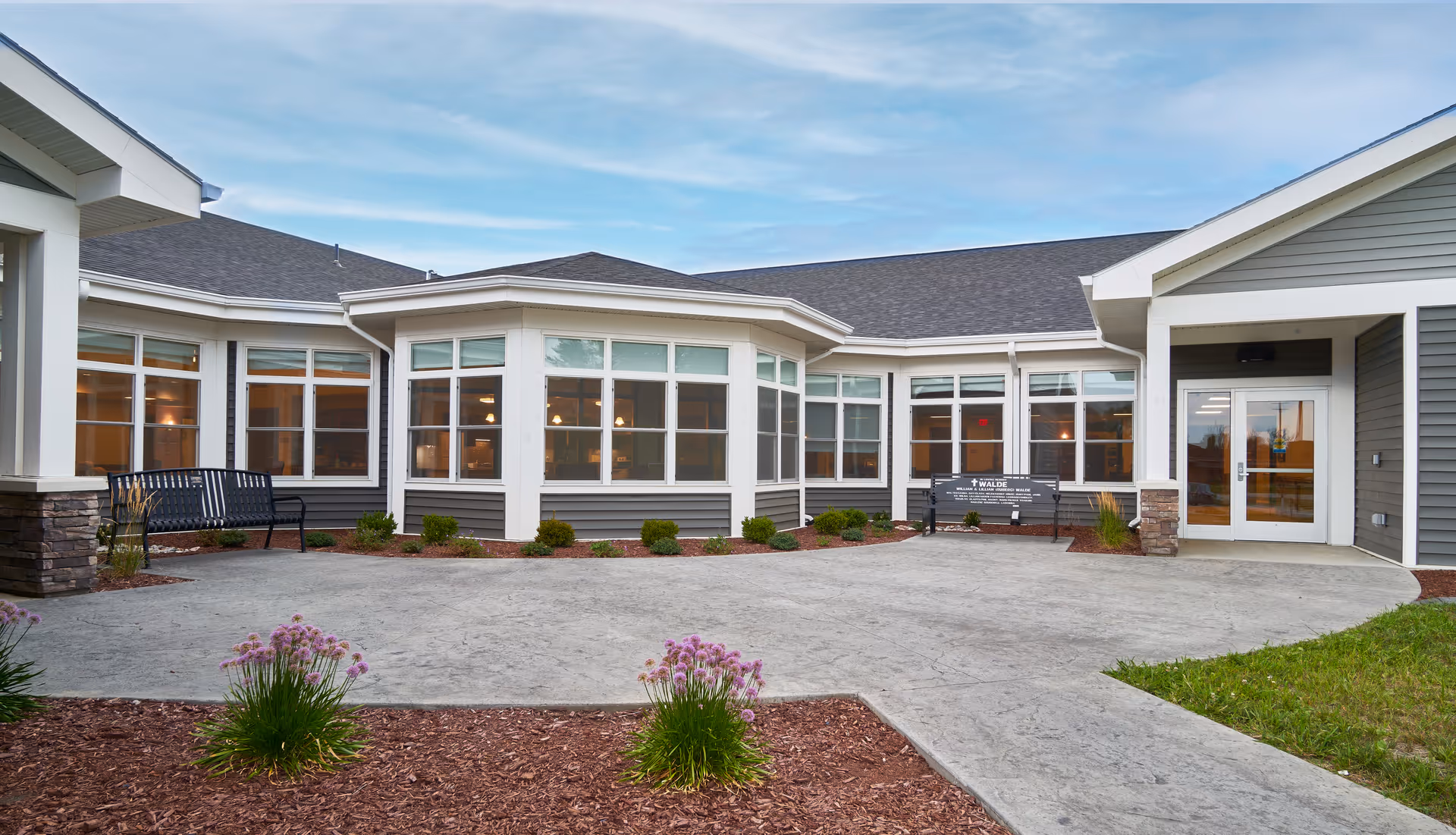 Exterior view of a single-story senior living facility building with large windows, a paved walkway, landscaped flower beds with purple flowers, and two black benches near the entrance under a partly cloudy sky.