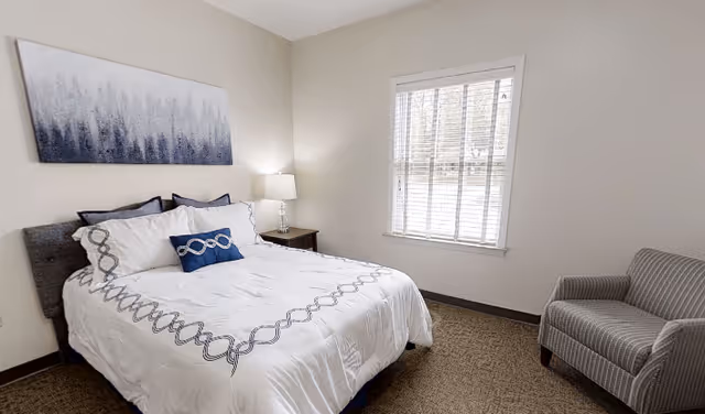 A neatly made bed with white and blue bedding in a simple bedroom. There is a nightstand with a lamp next to the bed, a window with blinds letting in natural light, a gray upholstered armchair in the corner, and a blue and white abstract painting on the wall above the bed.