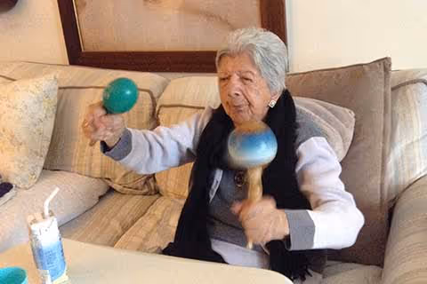 An elderly woman sitting on a beige couch in a living room, holding and shaking two maracas, one green and one blue.