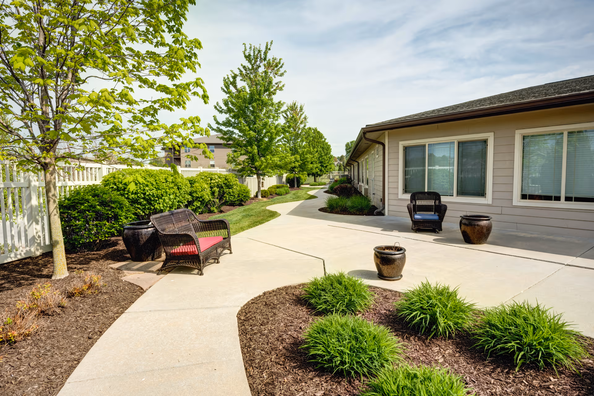 Outdoor patio area at Cedar Creek of Prairie Meadows featuring a paved walkway, green shrubs, small trees, and wicker chairs with cushions. The area is adjacent to a building with large windows and has several decorative pots placed along the walkway.