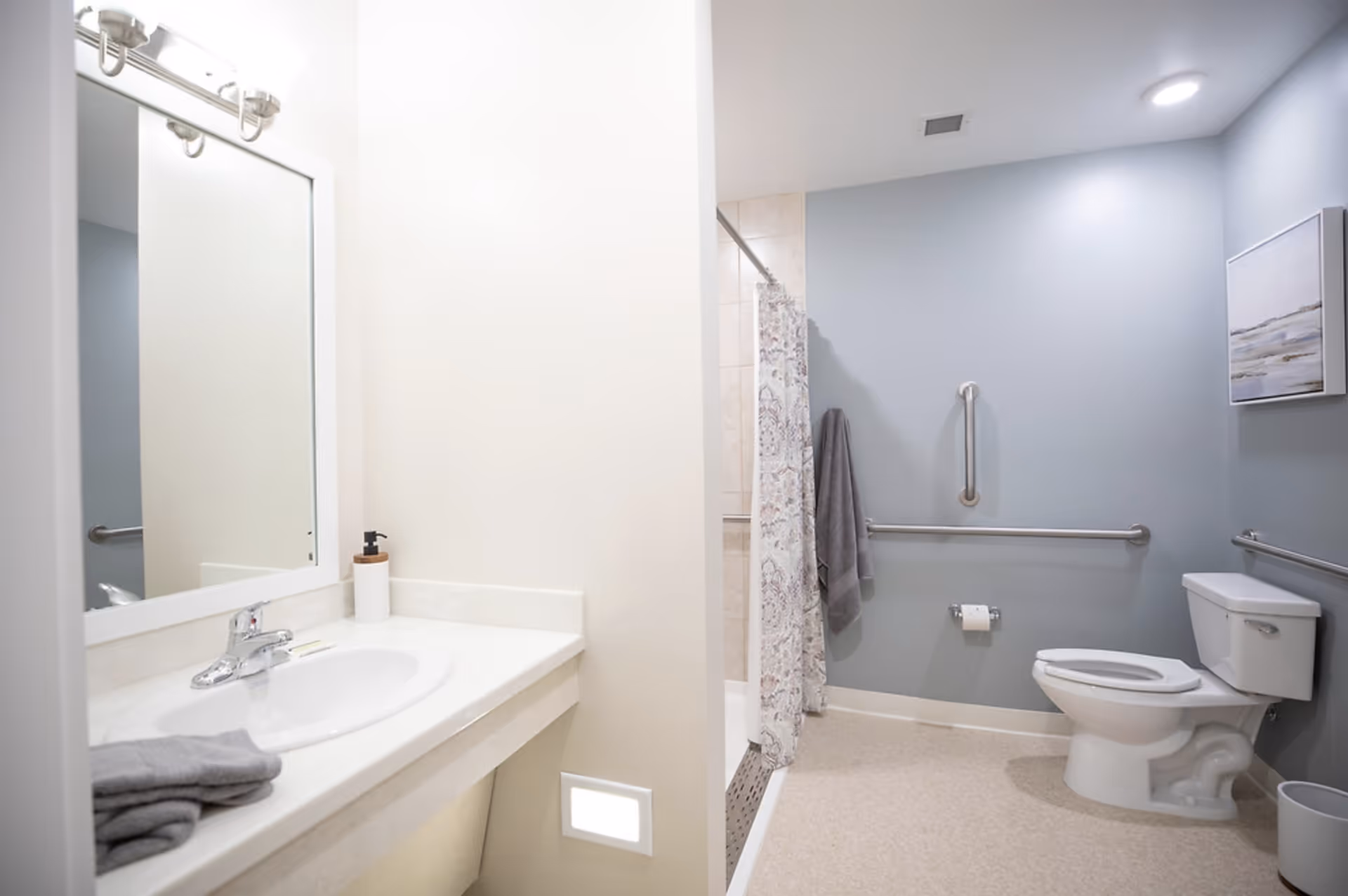 A clean and accessible bathroom with a white sink and countertop on the left, a large mirror above the sink, and a soap dispenser. On the right side, there is a toilet with grab bars on the walls for support, a toilet paper holder, a towel hanging on a hook, a shower area with a patterned curtain, and a framed picture on the wall. The walls are painted light blue and white, and the floor is light-colored.