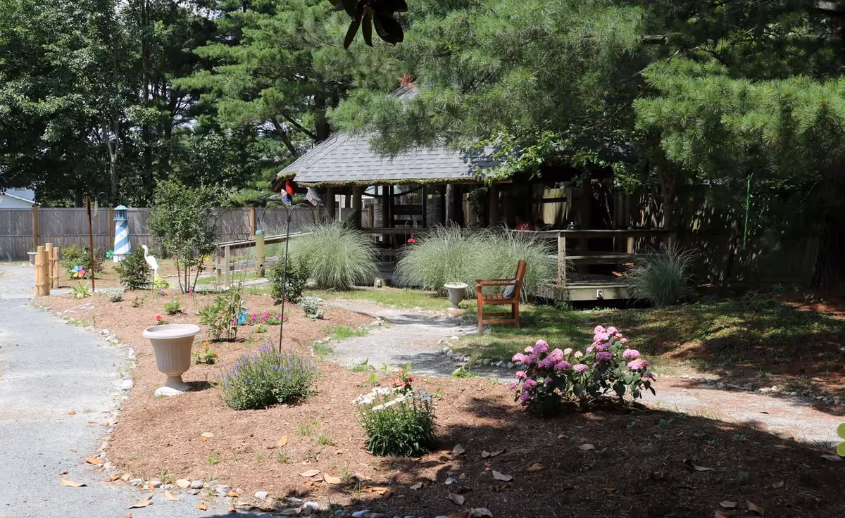 A peaceful outdoor garden area with a gravel pathway winding through flower beds containing various plants and flowers. There is a wooden bench along the path and a gazebo structure with a shingled roof surrounded by greenery and trees. A wooden fence encloses the area.