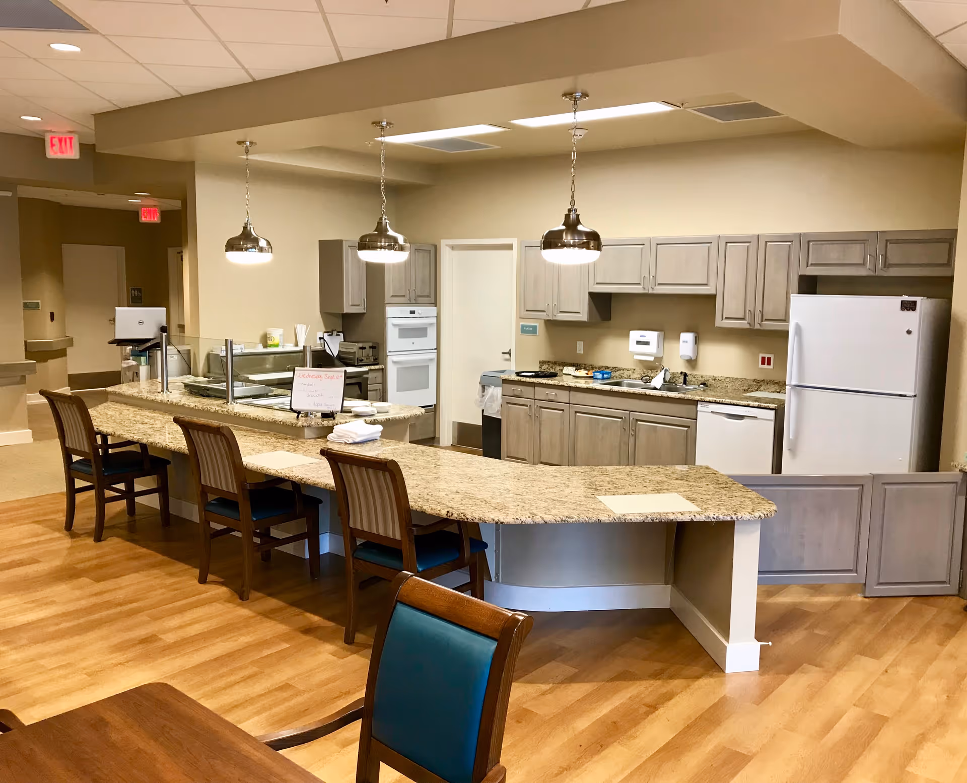 A modern kitchen area in a senior living facility with a long granite countertop bar featuring three wooden chairs. The kitchen has light wood cabinets, a white refrigerator, dishwasher, built-in oven, and a sink. Three pendant lights hang above the countertop. The floor is wood, and there are exit signs visible in the background.