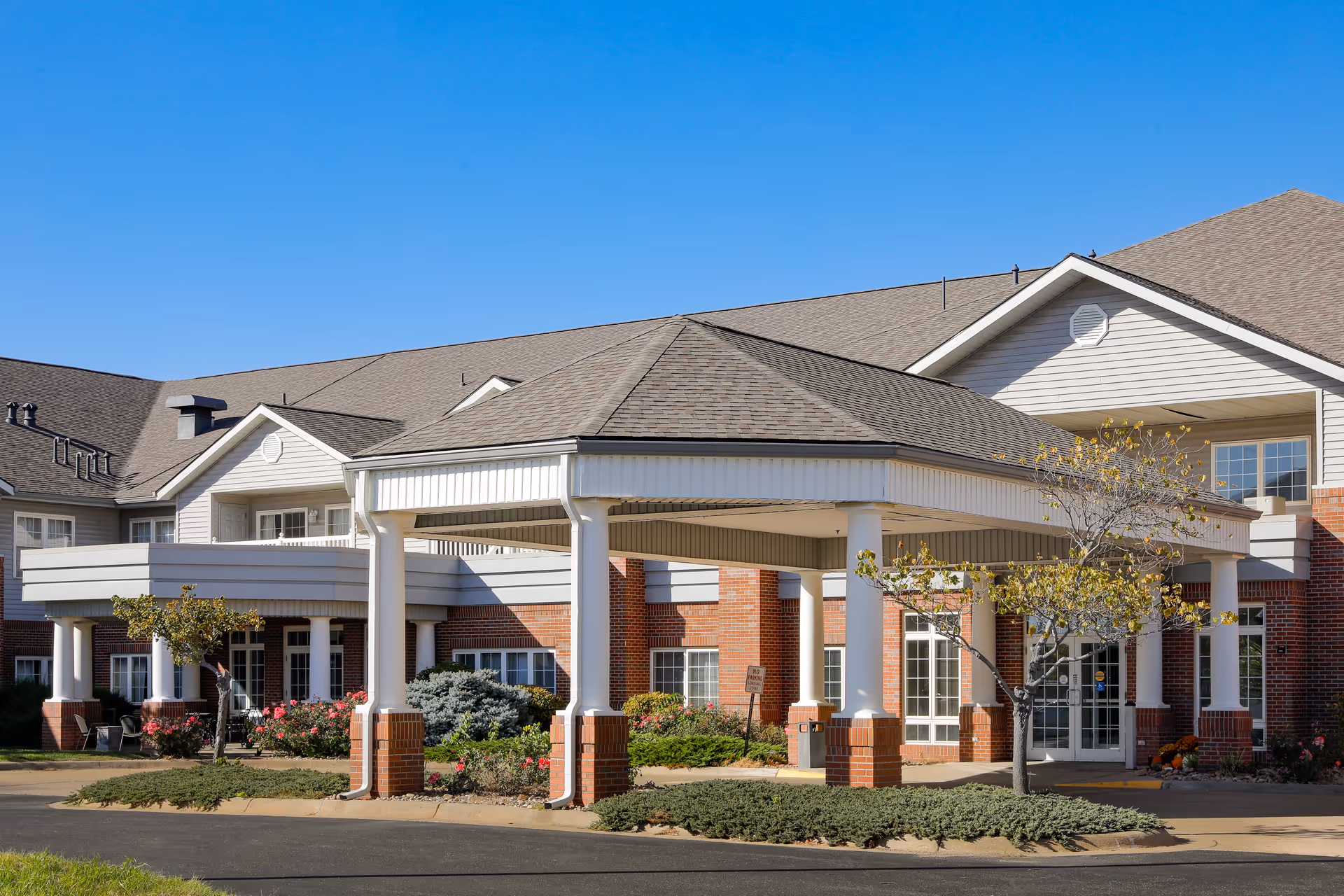 Exterior view of a senior living facility building with a covered entrance supported by white columns. The building has a combination of red brick and light-colored siding with multiple windows and a clear blue sky above.